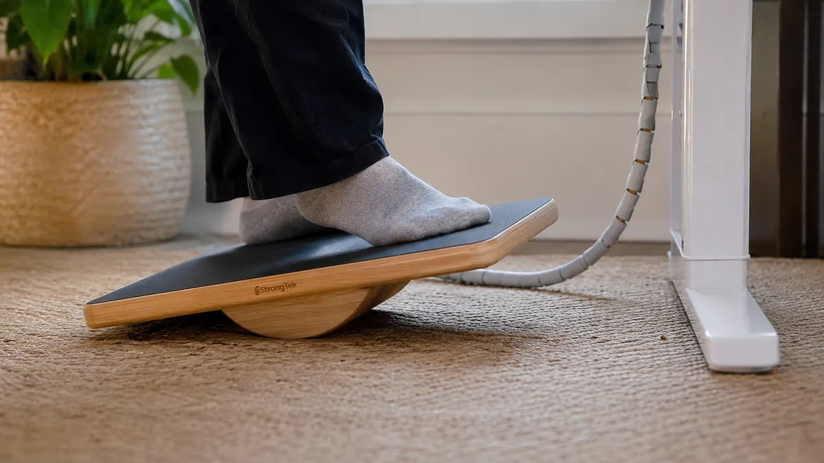 Close-up of a foot balancing on the StrongTek wooden wobble board next to a standing desk leg