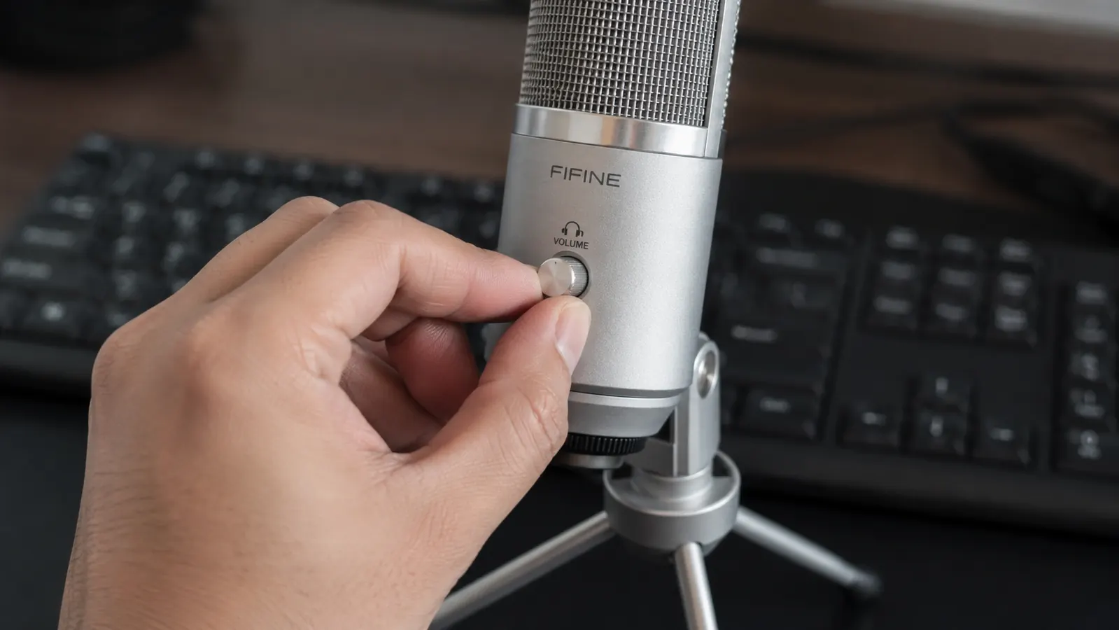 Hand adjusting the gain control knob on a silver metal desktop USB microphone next to a laptop keyboard